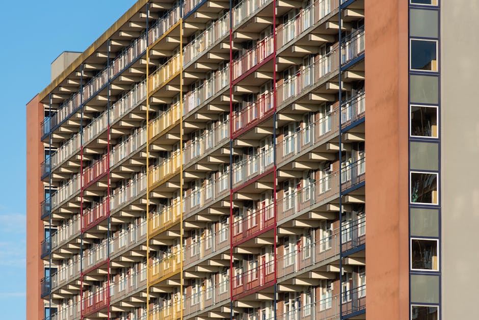 A high-rise residential building with multiple floors featuring balconies with metal railings painted in alternating colors of red, yellow, and blue. Each balcony has glass sliding doors leading into individual apartment units, with some open or partially visible through the glass. The building's exterior walls are painted in a muted beige and terracotta tone, and the right side has a vertical row of large, rectangular windows. The image is taken during daylight with a clear blue sky in the background. Although the primary focus is on the building's facade, the photograph's context hints at urban living, which can be relevant in house removals and furniture transport discussions, such as moving furniture out of apartments during home relocation services provided by Man with Van Turnpike Lane.