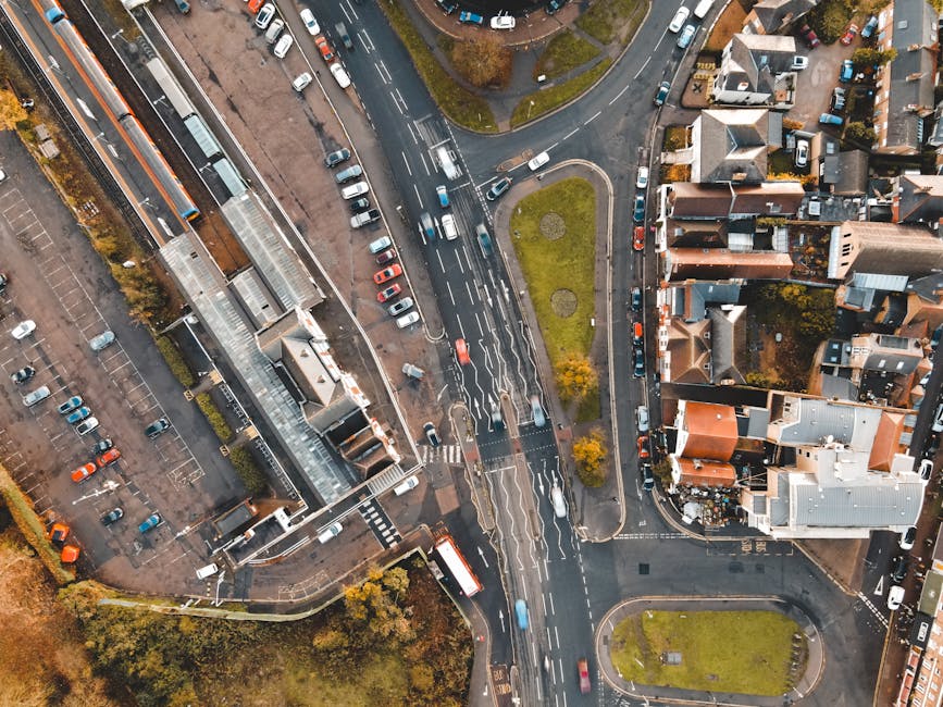 An aerial black-and-white photograph of a residential area showing terraced houses with pitched roofs closely arranged in rows along narrow streets. Behind the houses, there are numerous trees providing some greenery amid the tightly packed dwellings. In the foreground, a wide road with parked cars runs horizontally across the image, featuring a row of vehicles along the curb. The houses have small front gardens and some are separated by fences or walls. This scene captures a typical suburban neighbourhood, with the rooftops and streets clearly visible, illustrating the environment where home relocation and furniture transport services by Man with Van Turnpike Lane could be applicable during a house or flat move on Green Lanes Estate, highlighting the logistical context of packing and moving activities.