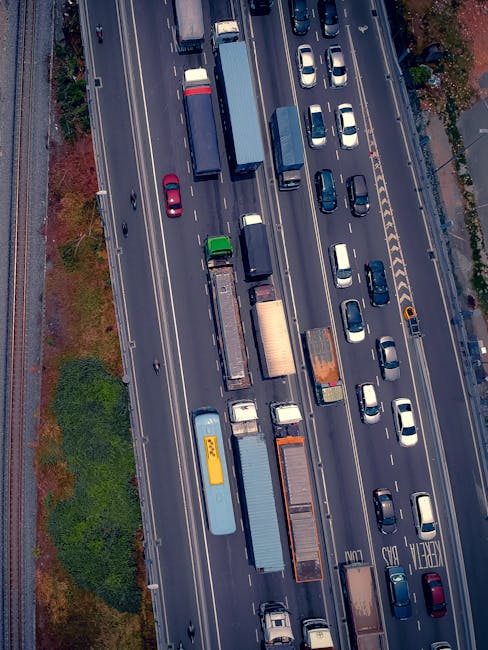 An aerial view of a busy multilane road showing several parked and moving vehicles, including trucks, vans, and passenger cars. On the left side, there is a patch of green trees and shrubbery adjacent to the roadside. The vehicles are aligned in close proximity, with some stationary and others in motion, reflecting typical urban traffic conditions. The scene captures a section of the road during daylight with clear weather, and the road markings are visible, indicating lanes and directional arrows. This image relates to the context of home relocation or furniture transport, highlighting the importance of effective vehicle and traffic management during removals services provided by Man with Van Turnpike Lane, especially for navigating busy areas such as Turnpike Lane in Haringey.