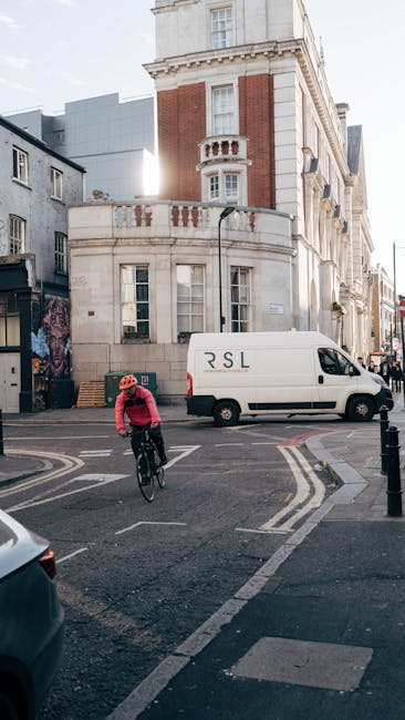 An urban street scene showing a cyclist wearing a red helmet and pink jacket riding a black bicycle across a marked pedestrian crossing at a corner in Turnpike Lane. To the right, a white delivery van with minimal branding is parked close to the curb, with its side door facing the street. Behind the van, there is a large white and red brick building featuring traditional architectural details, including a curved balcony with ornamental balustrades and tall windows. The building's upper floors incorporate a mix of stone and brickwork, with some modern extensions visible in the background. The street is illuminated by daylight, with shadows cast on the pavement, and the area appears to be part of a busy neighbourhood suitable for house removals and home relocation services operated by Man with Van Turnpike Lane. The scene captures elements relevant to furniture transport, loading areas, and urban logistics for removals.