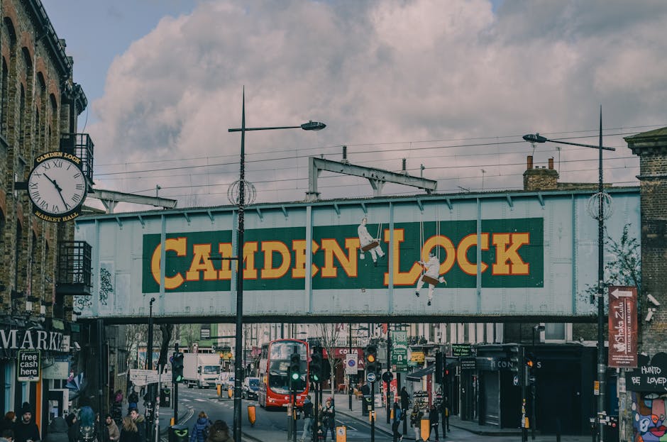 A view of Camden Lock in London featuring a large railway bridge with the words 'CAMDEN LOCK' painted in bold, yellow letters against a green background. The bridge is supported by metal beams and has