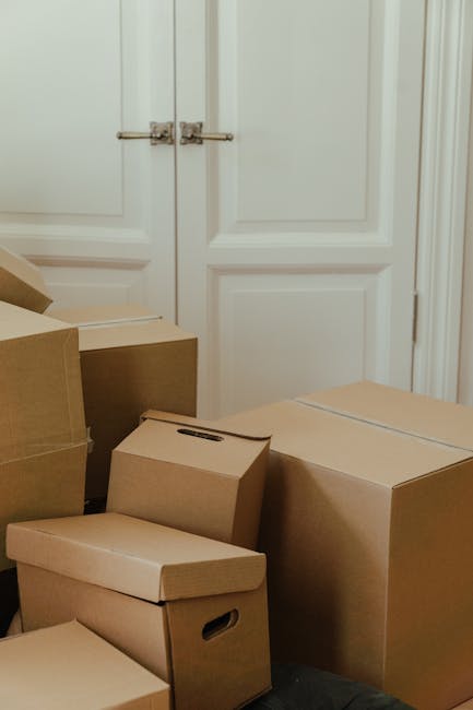 A collection of various cardboard moving boxes in different sizes, some with built-in handles, are stacked and placed on the floor near a closed white wooden door with decorative paneling and metallic hinges. The boxes appear to be ready for a home relocation or packing process, with some stacked on top of each other and others positioned side by side, indicating an ongoing moving preparation. The doorway and surrounding area suggest an indoor environment, possibly a hallway or room, with neutral lighting. This setup reflects the packing and moving stages typical of furniture transport and house removals services provided by companies like Man with Van Turnpike Lane, emphasizing organization and care during a house move.