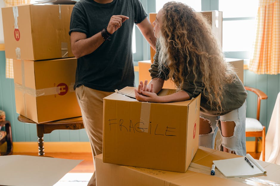 A woman with long, curly hair leaning over a cardboard moving box labeled 'FRAGILE' in handwritten text, inside a room with teal walls and checked yellow-orange curtains. She is engaging with a man, who is partially visible and wearing a dark t-shirt, as they prepare for home relocation. The scene shows several packed cardboard boxes stacked on a small wooden table and on the floor, with some wrapped in plastic or packing tape. A clipboard with a pen rests on the table next to the boxes, indicating packing or inventory planning. Bright natural light fills the room, highlighting the detailed packing process involved in furniture transport and moving logistics. Man with Van Turnpike Lane, a professional removals service, is preparing for a safe and efficient house move, ensuring proper handling of fragile items during the loading process.