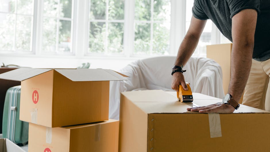 A collection of various cardboard moving boxes in different sizes, some with built-in handles, are stacked and placed on the floor near a closed white wooden door with decorative paneling and metallic hinges. The boxes appear to be ready for a home relocation or packing process, with some stacked on top of each other and others positioned side by side, indicating an ongoing moving preparation. The doorway and surrounding area suggest an indoor environment, possibly a hallway or room, with neutral lighting. This setup reflects the packing and moving stages typical of furniture transport and house removals services provided by companies like Man with Van Turnpike Lane, emphasizing organization and care during a house move.