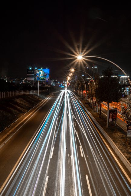 This image shows a multi-lane urban road at night captured with long exposure photography, where streaks of white and red light trail along the lanes indicating vehicle movement. The road is lined with streetlights, illuminated signs, and commercial billboards. The background features city buildings with lit windows, and the sky is dark. The scene highlights the busy traffic flow typical during home relocation or moving logistics, with the motion emphasizing the dynamic process of furniture transport and packing during house removals. The bright light streaks and urban environment reflect the urgency and activity involved in professional removals services such as those provided by Man with Van Turnpike Lane, especially when coordinating furniture transport within a city setting.