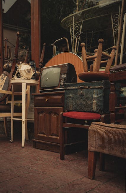 A collection of furniture and household items arranged on a paved outdoor surface, including wooden chairs with fabric and upholstered seats, small tables, a vintage television set, a cardboard box, and a blue patterned suitcase, with a metal garden table and a windowed building in the background. The furniture appears to be organized for packing or transportation, indicating a home relocation process. The scene is well-lit, capturing the details of the various objects as part of a furniture loading and packing process, consistent with a house removal service by Man with Van Turnpike Lane during a furniture move.