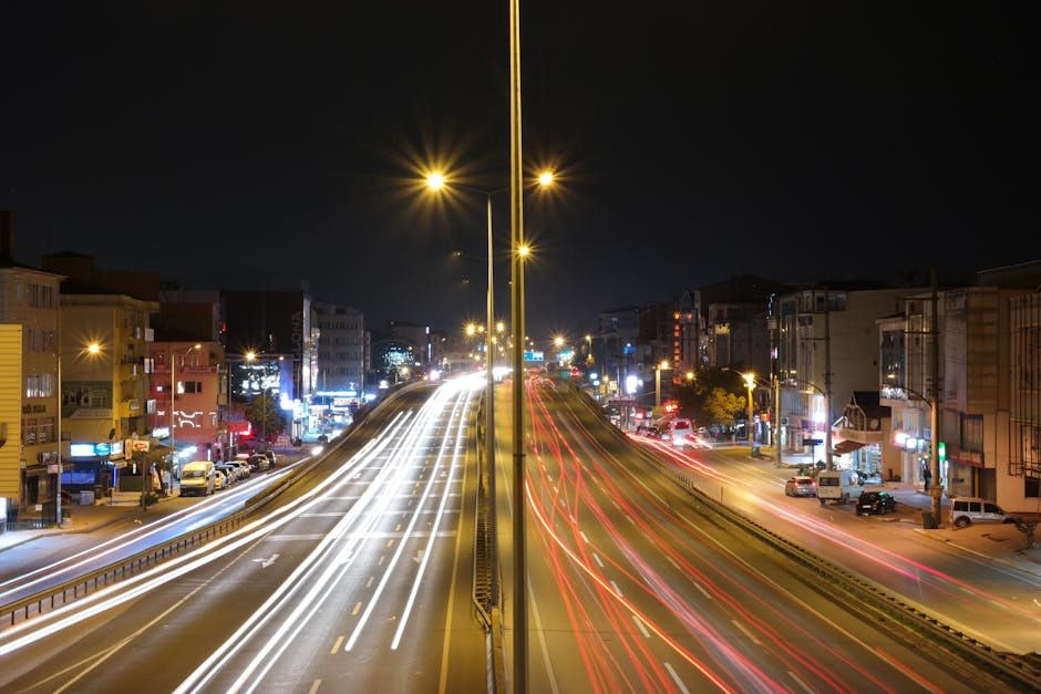A long-exposure photograph of a busy urban street at night, with multiple lanes for traffic separated by a central divider. Light trails from moving vehicles create white streaks on the left side, ind