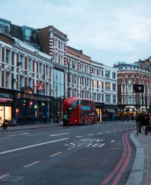 A street scene outside a row of multi-storey Victorian-style buildings with white and red brick facades, large windows, and decorative architectural details, illuminated by streetlights. In the foregr