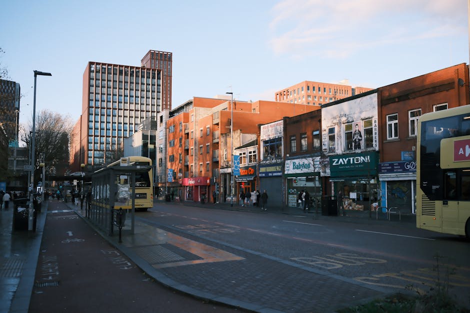 A street scene outside a row of multi-storey Victorian-style buildings with white and red brick facades, large windows, and decorative architectural details, illuminated by streetlights. In the foregr