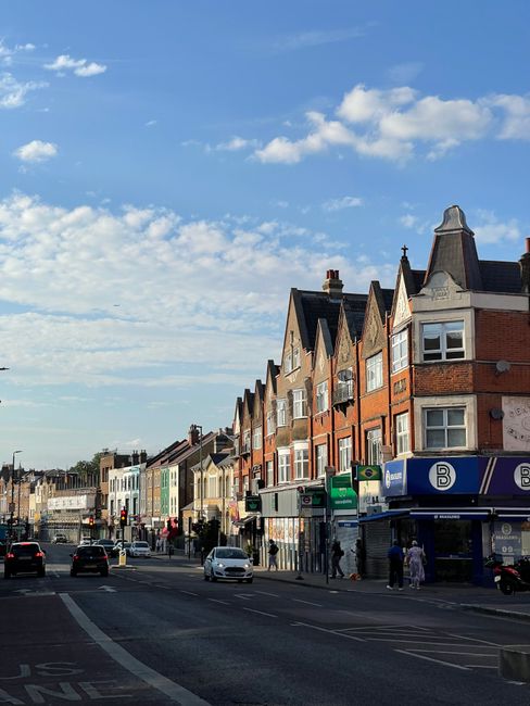 The image depicts a busy residential street in Hornsey, with a row of multi-storey brick houses featuring bay windows and gabled roofs. On the street, there are several parked cars and a few moving vehicles, with pedestrians walking along the pavement. In the foreground, a commercial unit with signage is visible, and the sky above is partly cloudy with patches of blue. This setting illustrates an urban environment suitable for house removals and furniture transport services, as provided by Man with Van Turnpike Lane. The scene captures the typical suburban street scene relevant to home relocation activities, with clear access for moving vehicles and packages, including cardboard boxes and packing materials possibly visible in new or current interiors or on the pavement during the loading process.