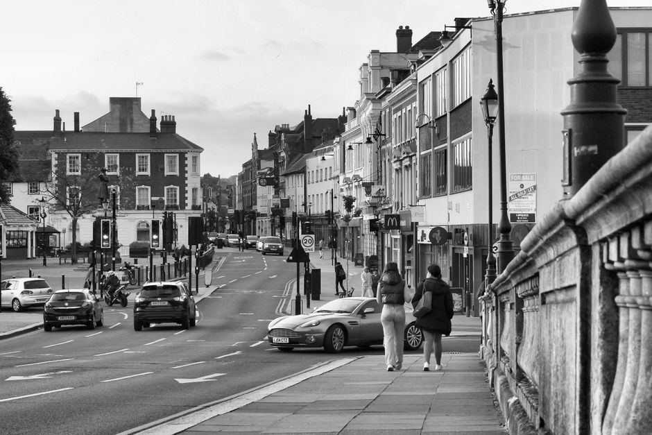 The image depicts a busy residential street in Hornsey, with a row of multi-storey brick houses featuring bay windows and gabled roofs. On the street, there are several parked cars and a few moving vehicles, with pedestrians walking along the pavement. In the foreground, a commercial unit with signage is visible, and the sky above is partly cloudy with patches of blue. This setting illustrates an urban environment suitable for house removals and furniture transport services, as provided by Man with Van Turnpike Lane. The scene captures the typical suburban street scene relevant to home relocation activities, with clear access for moving vehicles and packages, including cardboard boxes and packing materials possibly visible in new or current interiors or on the pavement during the loading process.