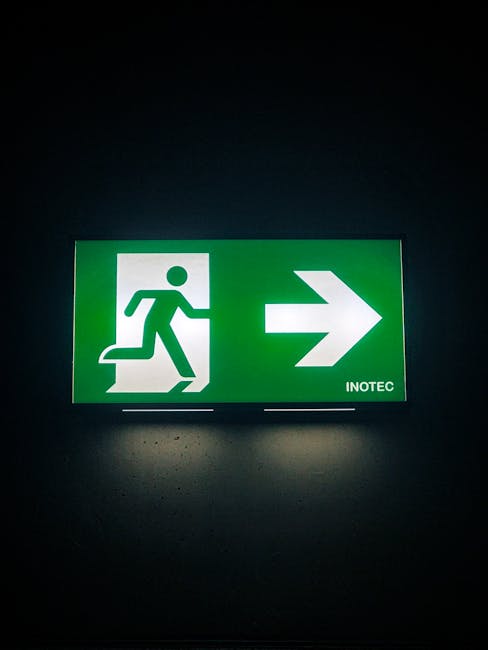 A green emergency exit sign with a white pictogram of a person running through a door, positioned on a dark wall. The sign features a large white arrow pointing to the right and the word 'INOTEC' in small text at the bottom right. The light from the sign illuminates the surrounding wall, highlighting its role as a directional guide for safe evacuation. This type of signage is commonly found in commercial or residential buildings, supporting safety and efficient movement during a house relocation or emergency scenario, as part of professional removals services by Man with Van Turnpike Lane.